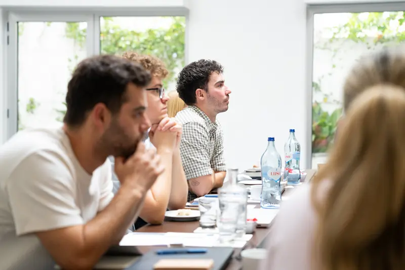 Team members listening along one side of a long table, garden windows behind them.