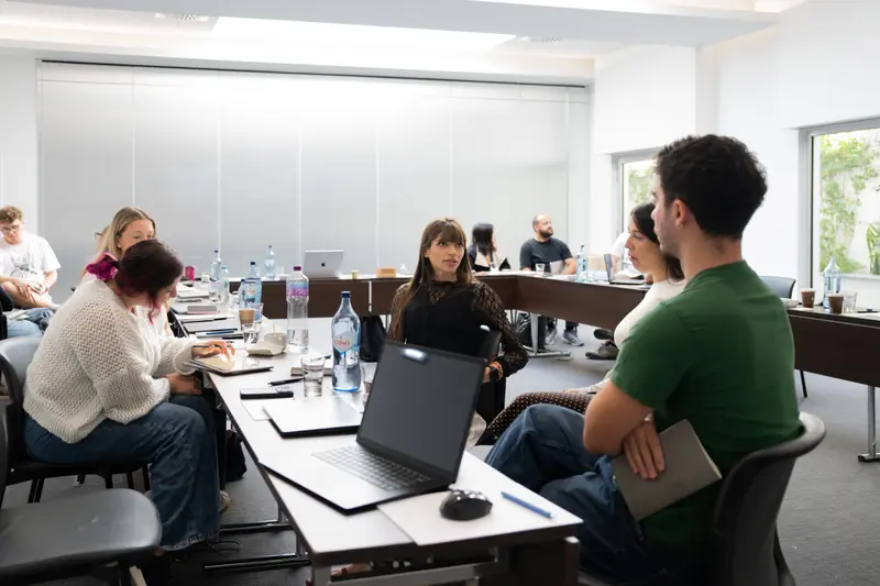 The wider room during a working session, small groups spread across tables with laptops and notes.