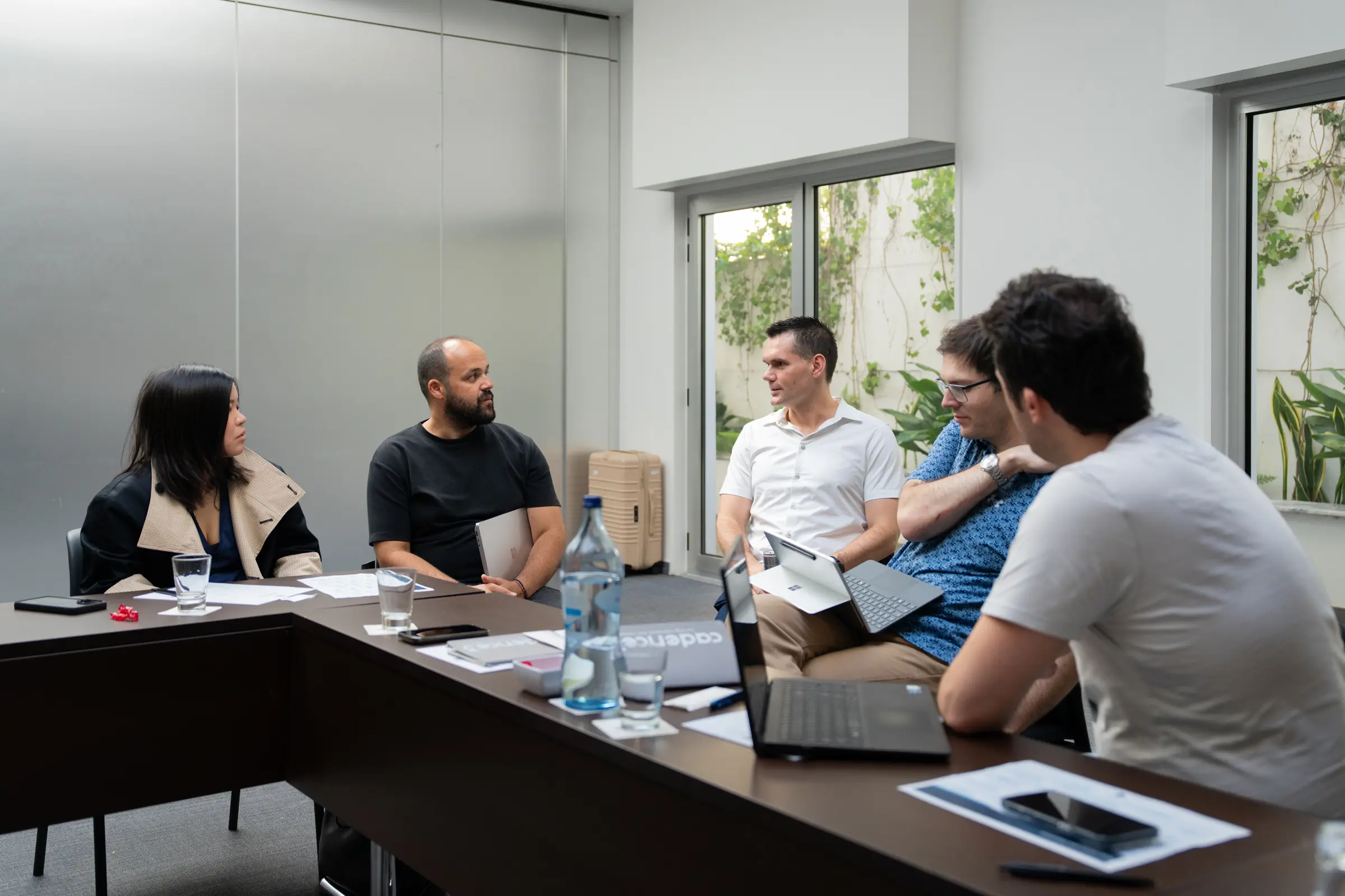 Six colleagues in conversation around a boardroom table, courtyard greenery through the window behind them.