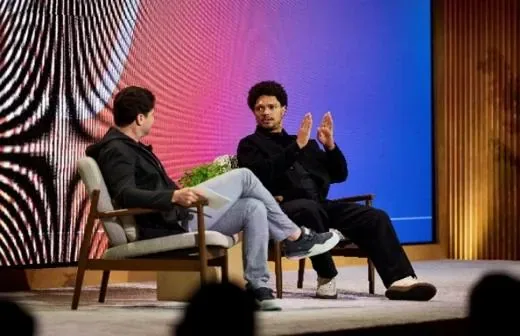 Two speakers seated on stage mid-discussion, a patterned LED backdrop behind them.