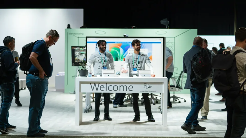 A green-walled Manufacturing stand with a 'Welcome' desk, attendees passing in front of it.