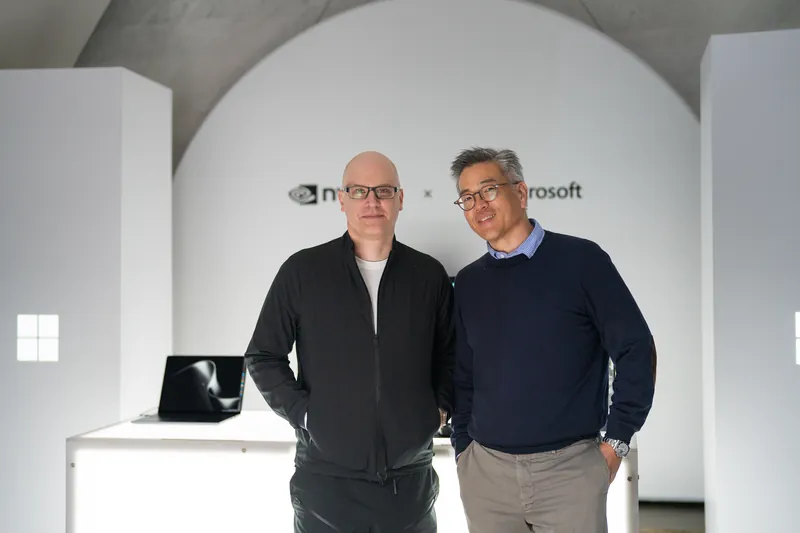 Two executives stand together in front of the NVIDIA and Microsoft wall, a laptop open on the plinth beside them.