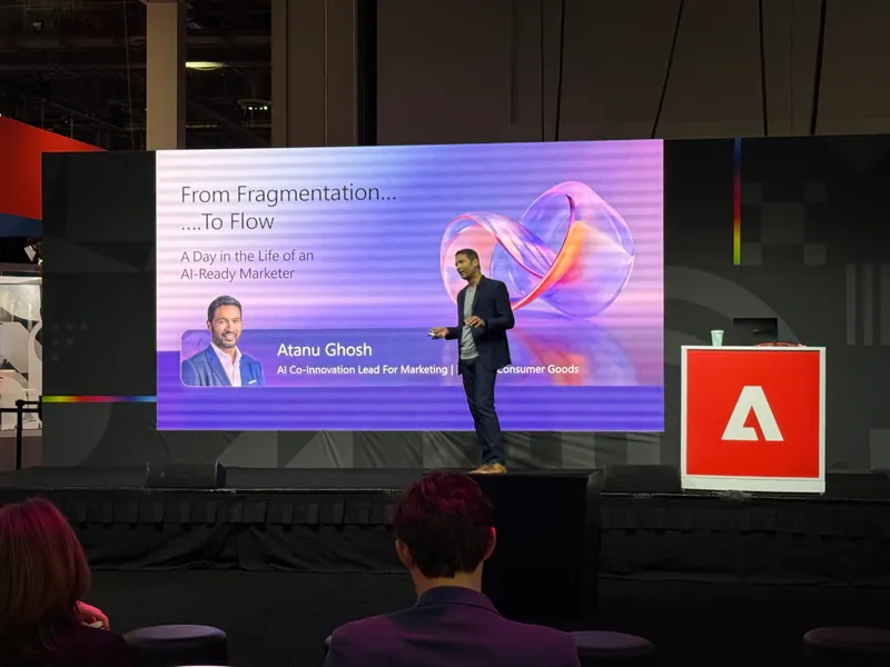 A speaker on stage in front of a screen reading “From Fragmentation To Flow”.