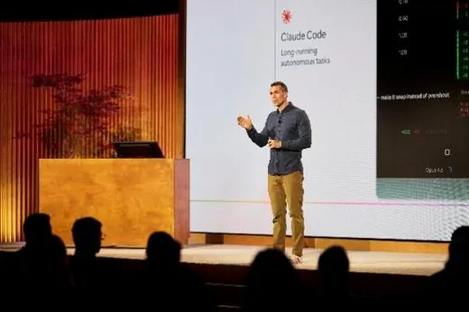 A speaker presenting to a seated audience, the session slide lit behind them.