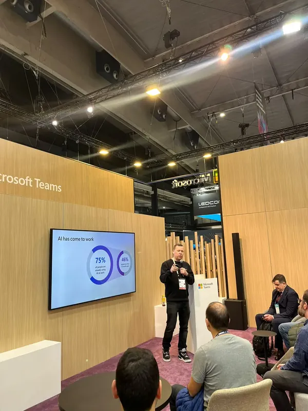 A Microsoft Teams speaker presenting workplace AI adoption figures to a seated audience in the stand theatre.